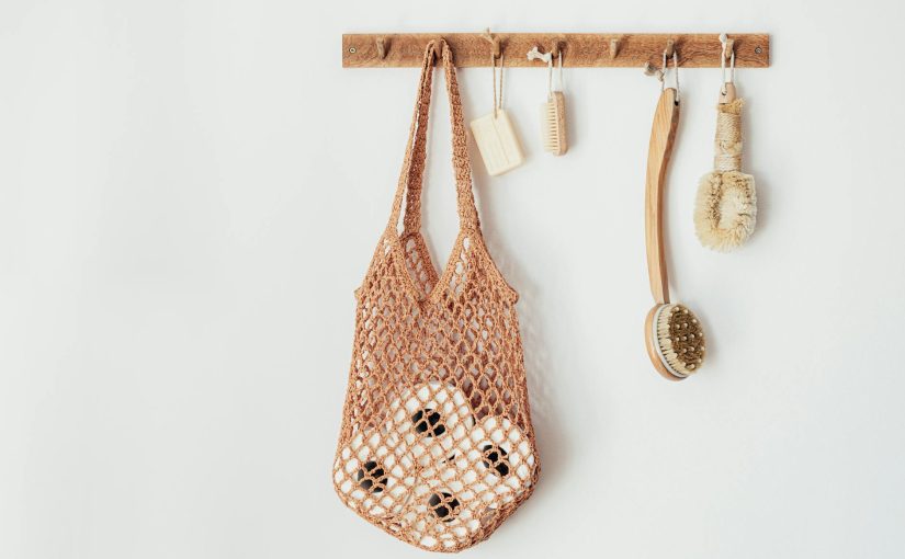 Composition with assorted wooden body brushes of different sizes with organic soap and string bag filled with toilet paper rolls hanging on wooden hanger on white wall in bathroom