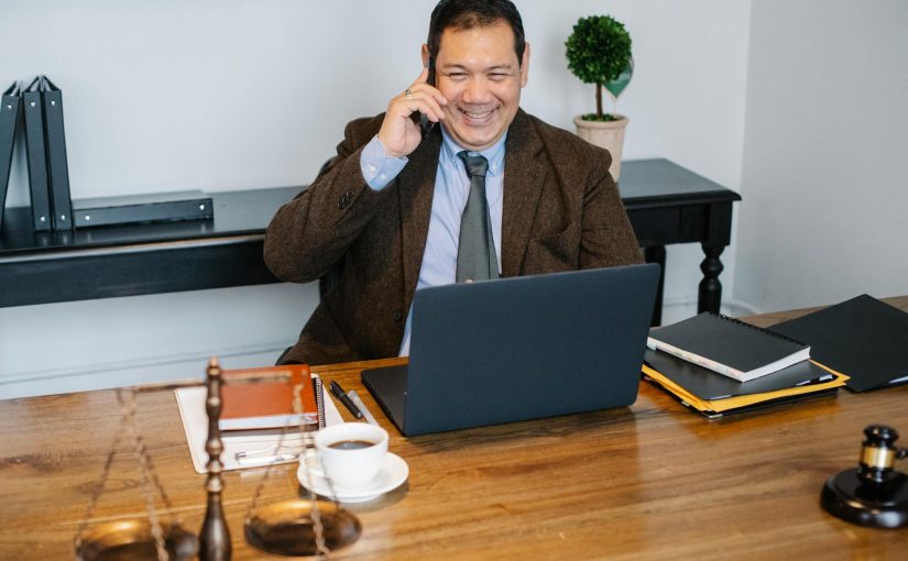 Joyful Asian male lawyer wearing formal suit having conversation on mobile phone and using laptop while working in modern law firm